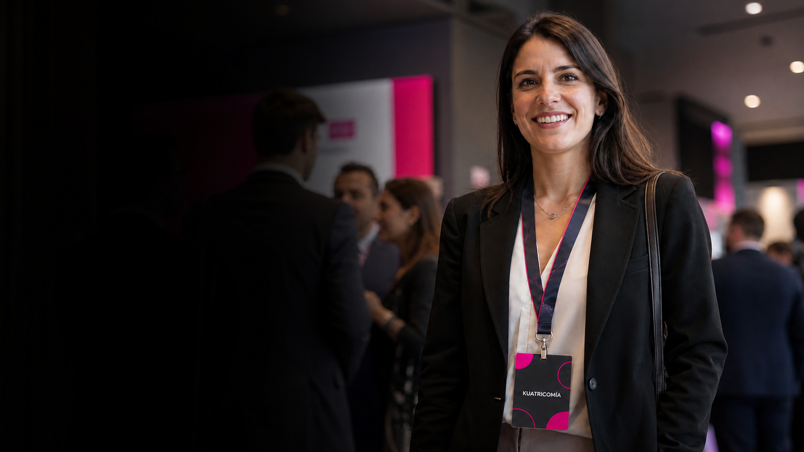 Smiling woman in a black blazer with a pink lanyard and conference badge at a networking event, background attendees blurred.